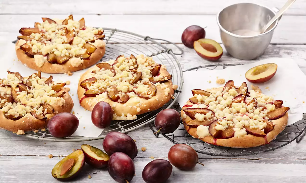 Okrugli kolači sa zlatno-smeđom podlogom, sočnim kriškama šljiva tamnoljubičaste boje i svijetlim mrvicama na žičanom stalku