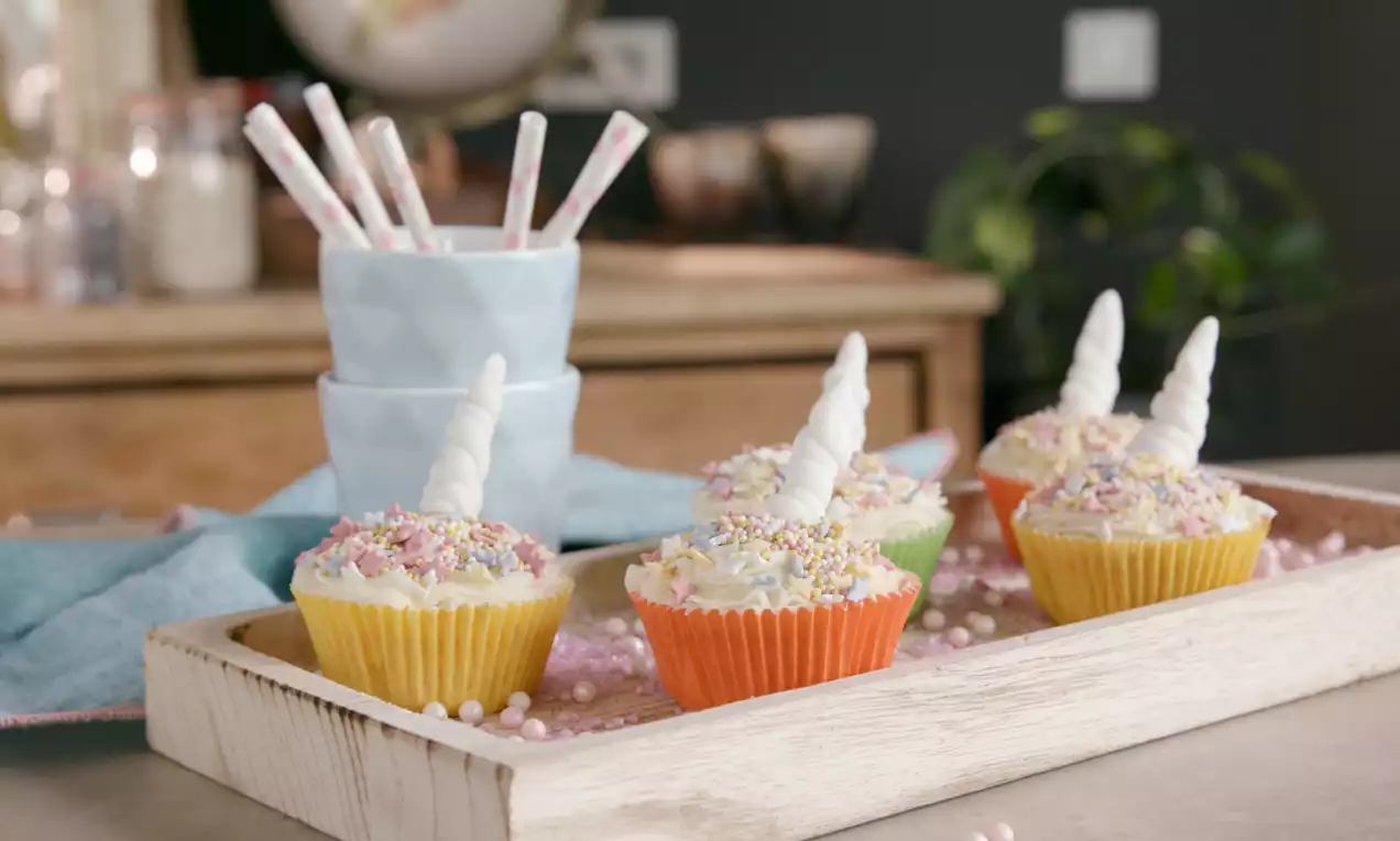Cupcakes colorés en caissettes jaune, orange et vert, garnis de crème blanche, cornes en sucre et éclats multicolores dans un plateau