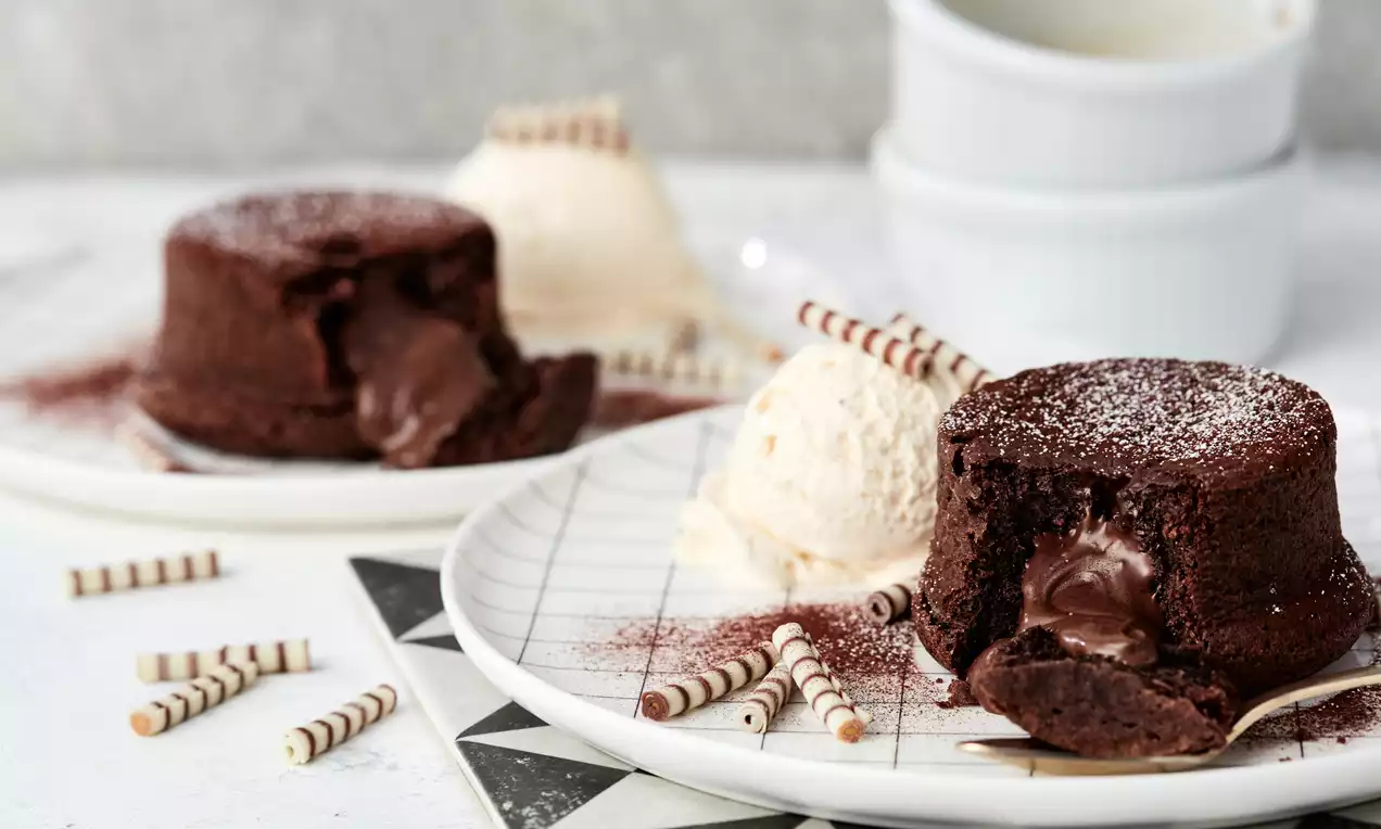 Schokoladen-Lava-Cake mit fließender Füllung auf weißem Teller, daneben Vanilleeis und gestreifte Dekorrollen, leicht mit Puderzucker bestäubt