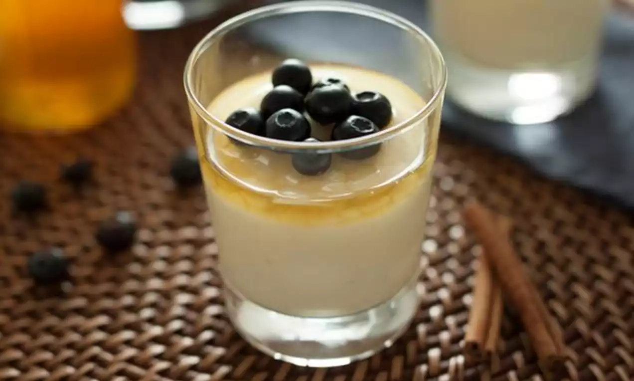 Pudim cremoso bege claro em copo transparente, coberto por calda caramelizada brilhante e mirtilos frescos, sobre mesa de fibra trançada