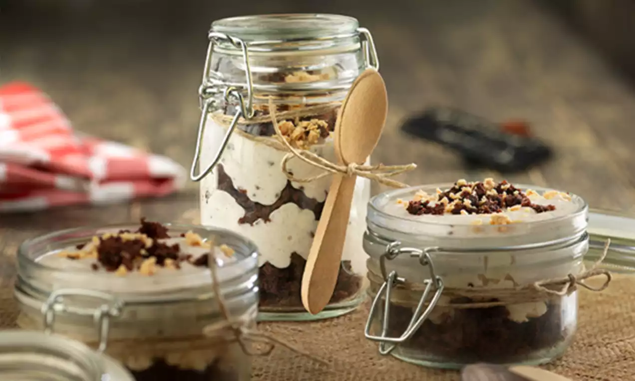 Postre en capas con crema beige clara y migas de bizcocho oscuro en frascos de vidrio con cierre metálico y cucharas de madera atadas