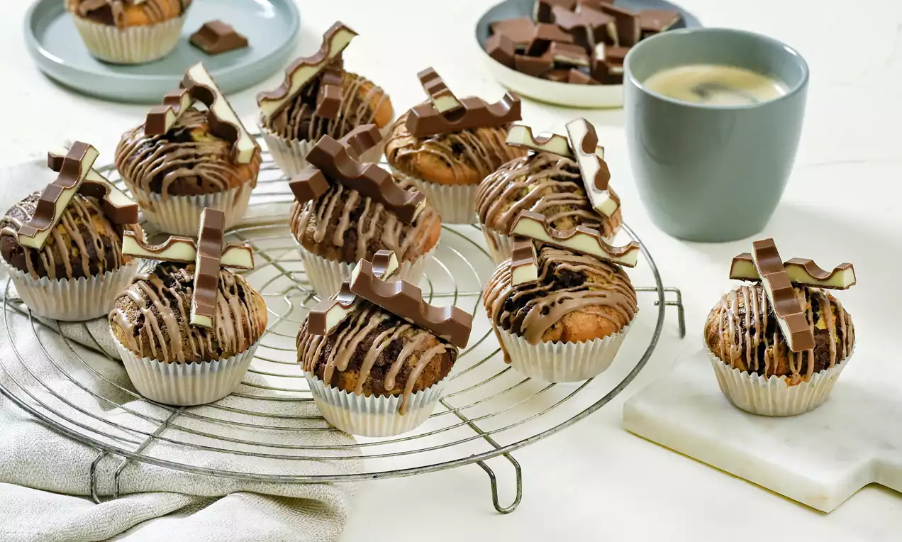 Kinder-Riegel®-Muffins mit goldbraunem Teig und Schokoglasur in weißen Förmchen, dekoriert mit Kinder-Riegel-Stücken auf einem Gitter