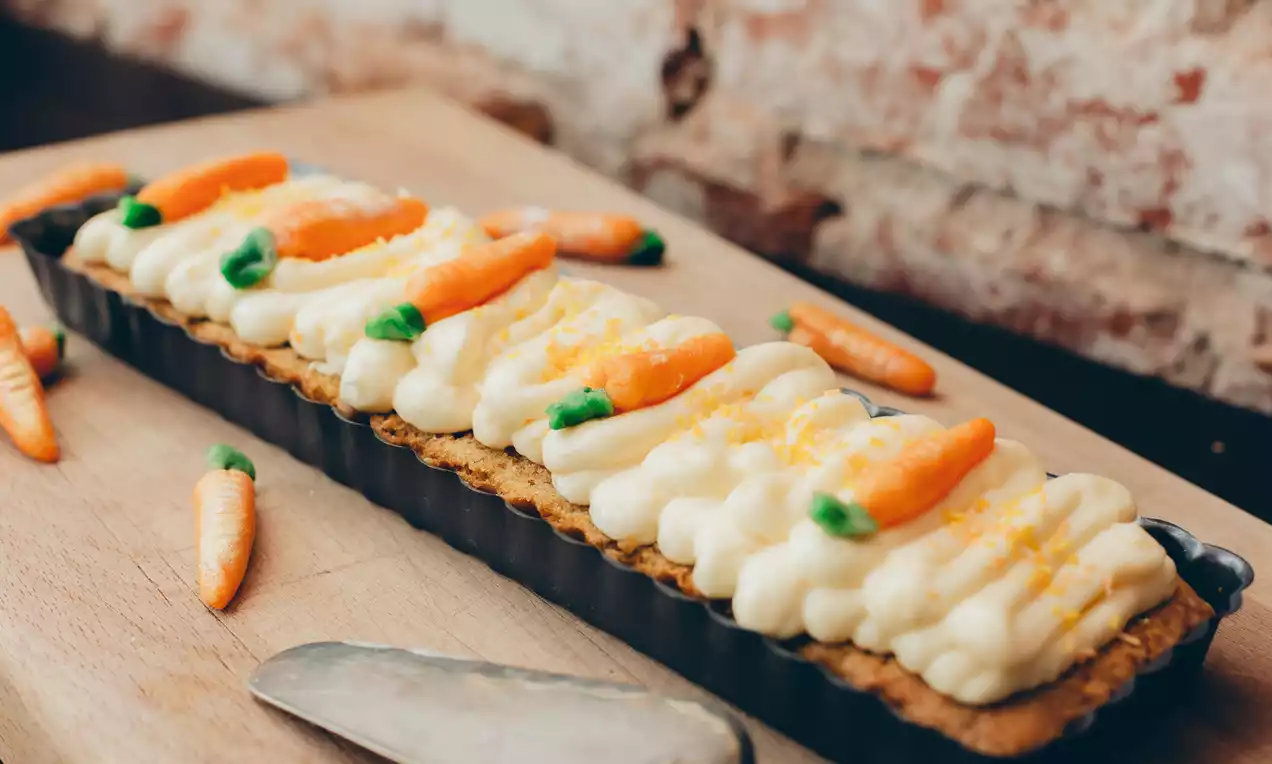 Pitkä suorakulmainen porkkanakakku vaalealla kremillä, koristeena oranssit ja vihreät marsipaaniporkkanat sekä hennosti raastettua appelsiininkuorta