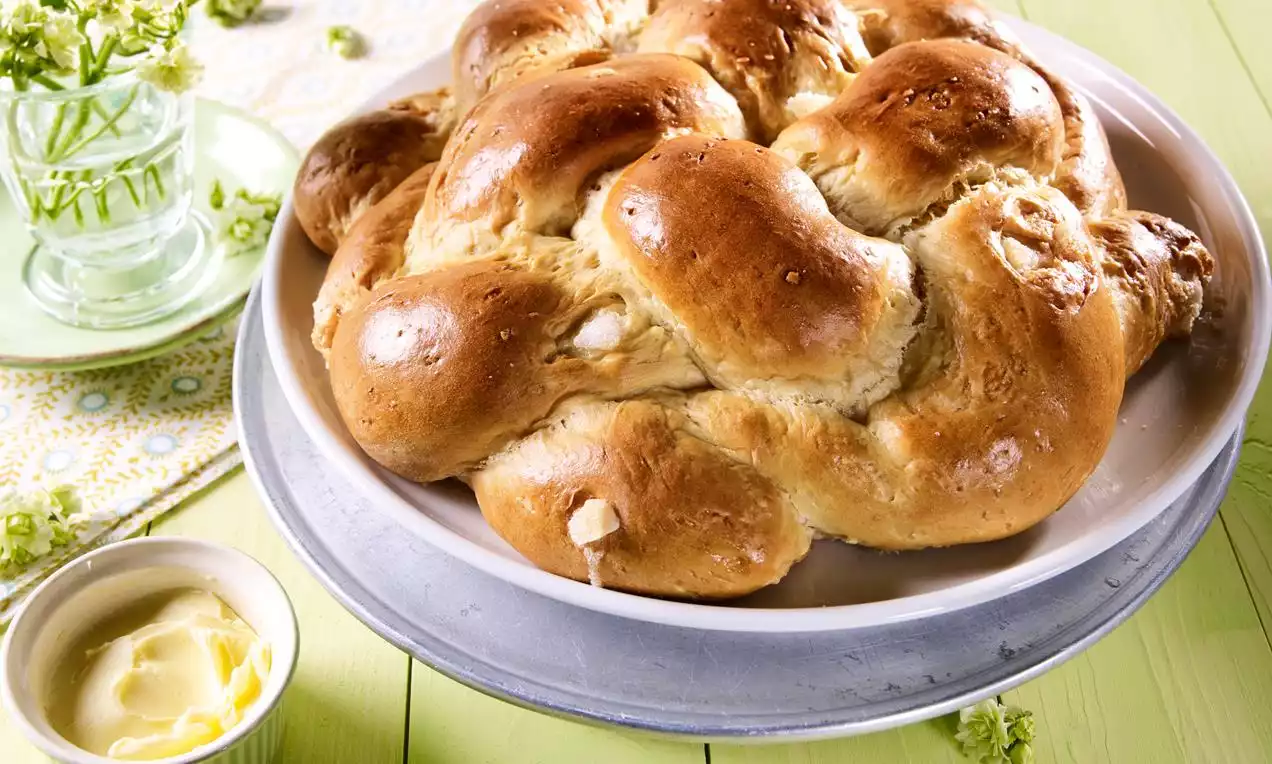 Aranybarnára sült, fényes, fonott kalács lágyan kidomborodó csomóival, amely egy világos tányéron pihen zöldes háttér előtt