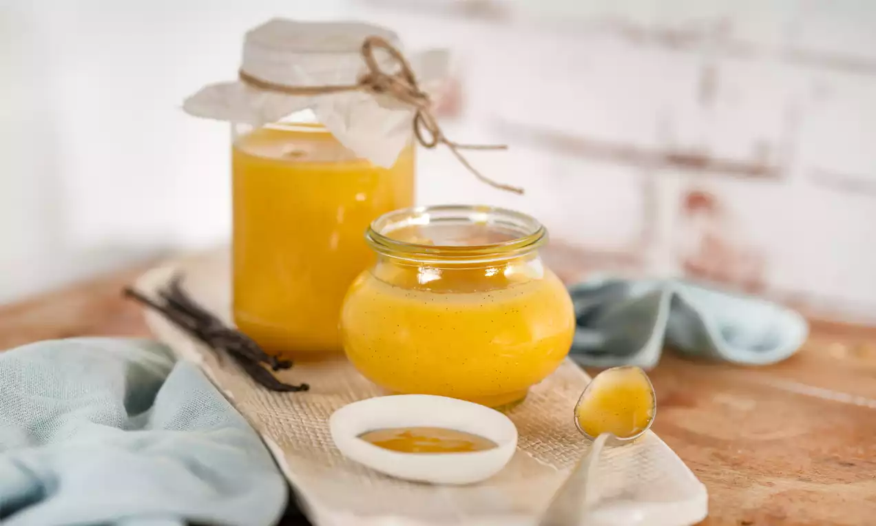Kleines Glas mit cremig-gelbem Vanille-Curd, daneben ein Löffel mit Curd und ein weißer Teller auf rustikalem Holzbrett mit Tuch drapiert
