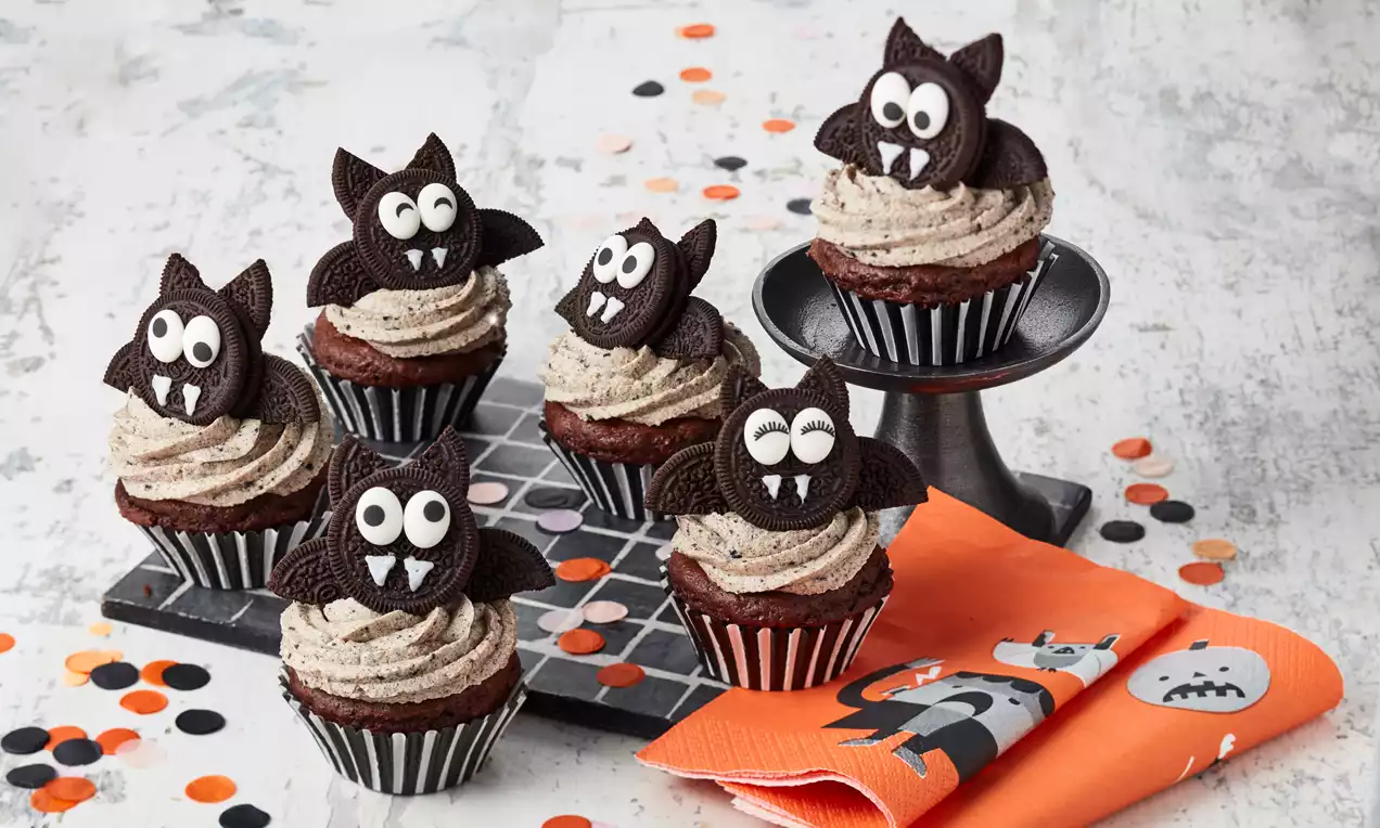 Schokoladen-Cupcakes mit grauer Cremehaube, dekoriert mit Oreo-Fledermäusen, großen Zuckeraugen und weißen Fangzähnen auf einem Tablett