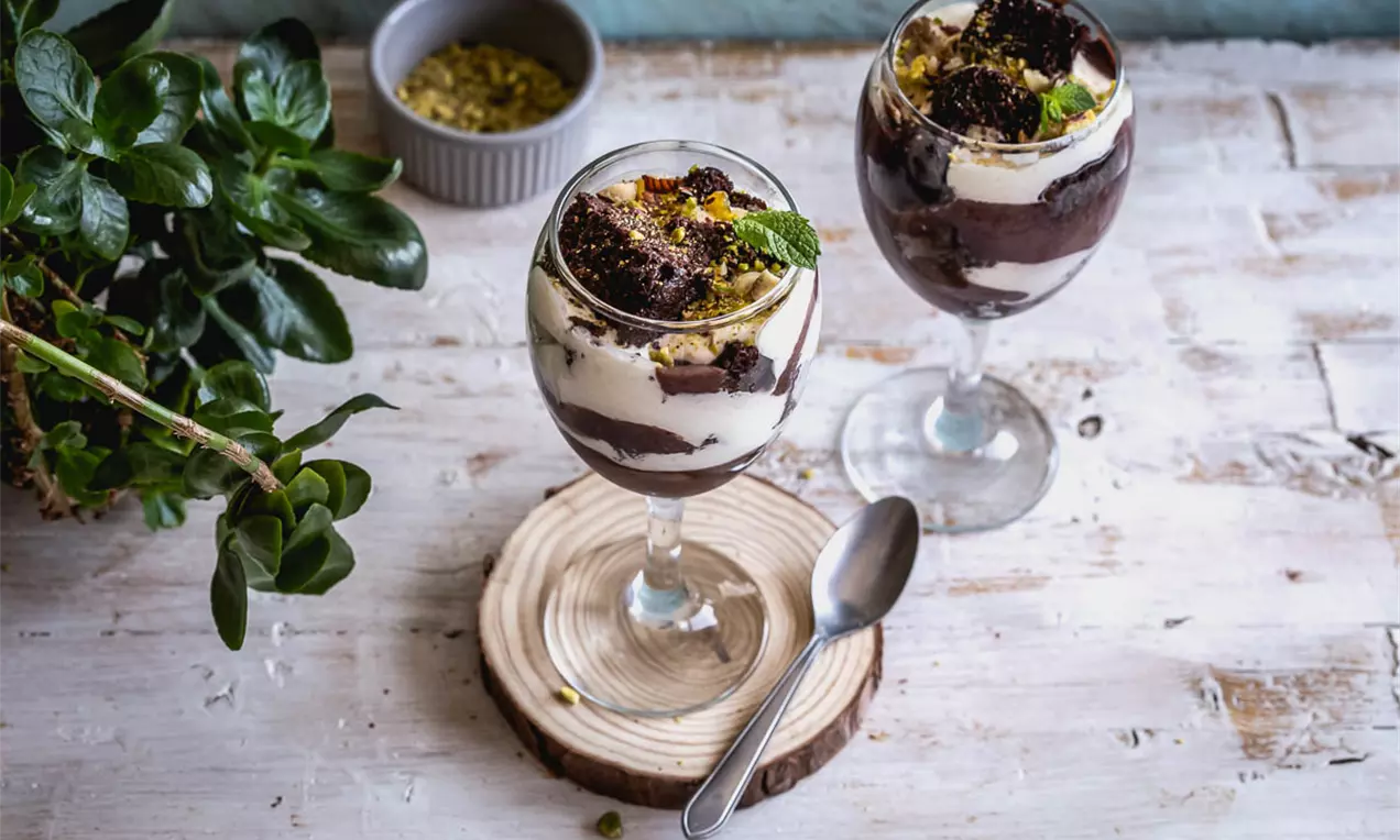 Слоеве от крем с ванилен нюанс и тъмнокафяв шоколадов кейк в стъклени чаши, гарнирани с натрошено шамфъстък и свежа зелена мента