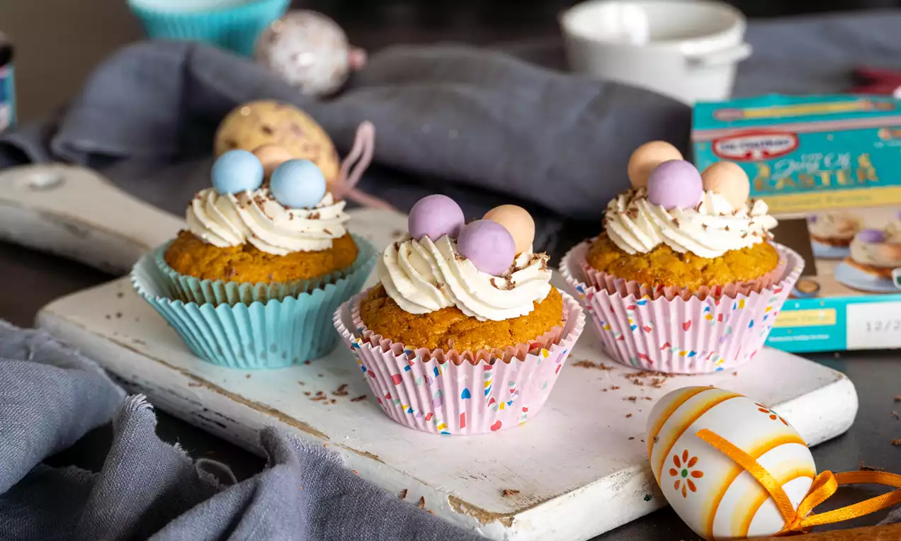 Muffini zlaćaste boje, u šarenim papirnatim košaricama, s kremastim bijelim vrhom i ukrasom od čokoladnih jaja pastelnih tonova