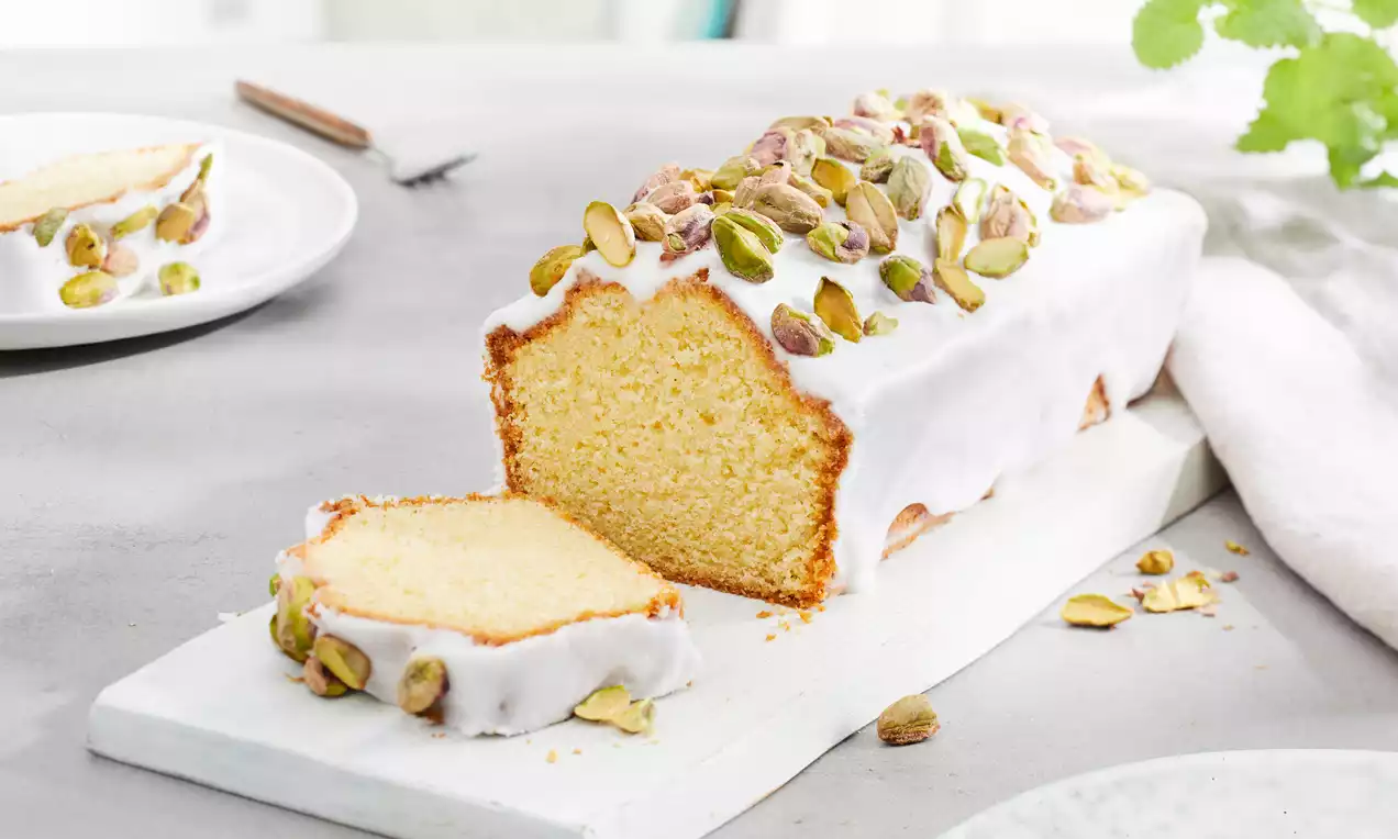 Golden loaf cake with white vanilla glaze topped with green pistachios, one slice cut and served on a white plate nearby