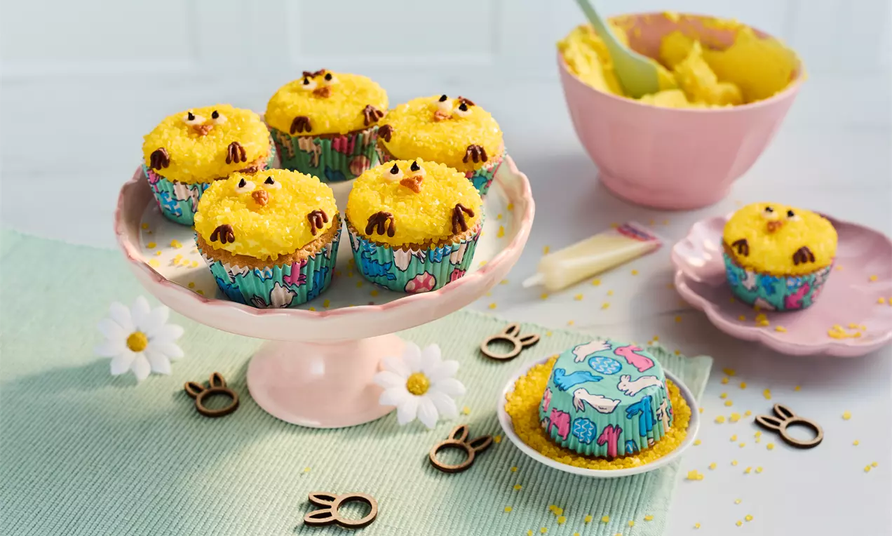 Yellow chick cupcakes with fluffy textured tops, chocolate details, arranged on a pink cake stand surrounded by spring-themed decor