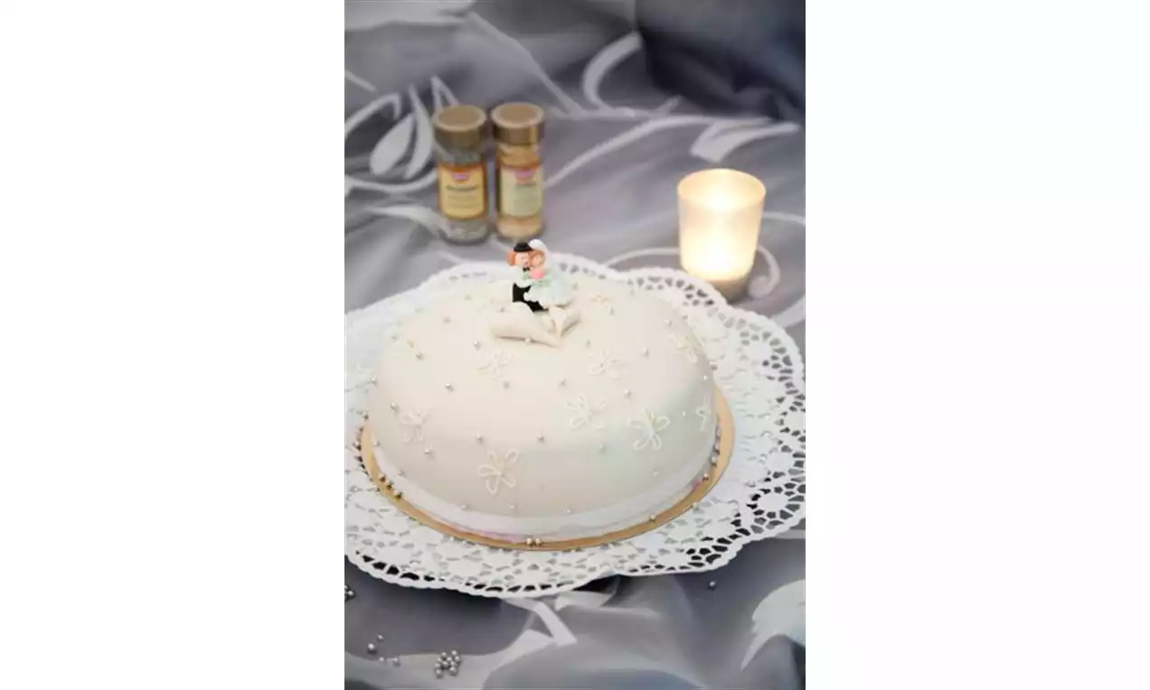 Rund hvit kake med perlemorsdekor og brudefigur på blondetallerken, omgitt av lys og krydderglass på grått mønstrete underlag