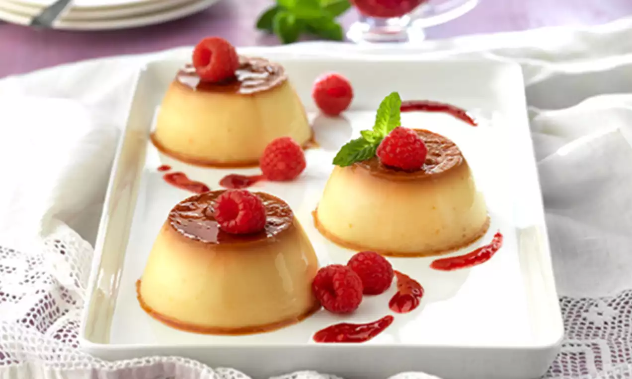 Flanes dorados de textura lisa con caramelo brillante, decorados con frambuesas frescas, hojas de menta y salsa roja sobre un plato blanco