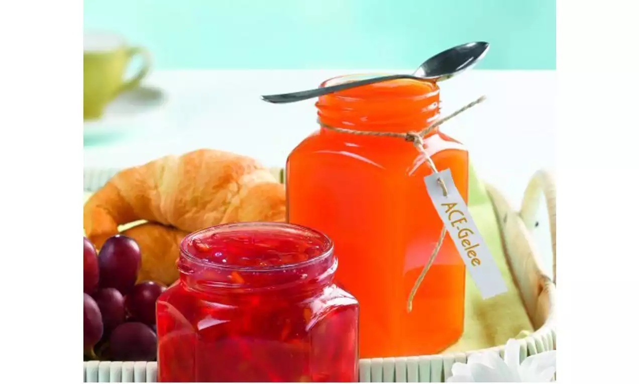 Oranges ACE-Gelee im Glas mit schwarzem Löffel auf einem geflochtenen Tablett neben roter Konfitüre, Croissant und violetten Trauben