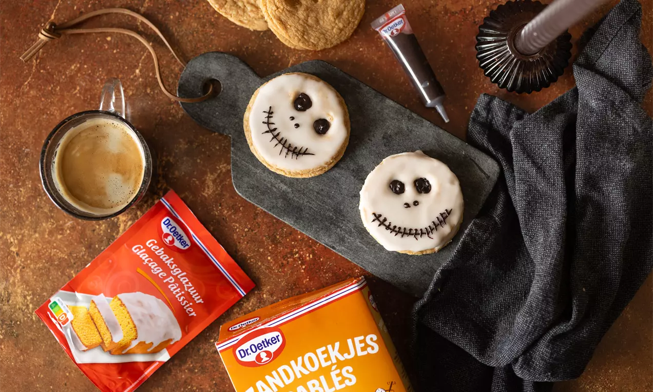 Zandkleurige ronde koekjes met witte glazuurlaag en zwarte mummiegezichten, liggend op een donker bord naast zwarte en rode decoratie