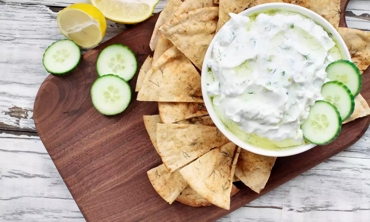A white bowl of creamy tzatziki with green cucumber pieces and dill, surrounded by golden pita triangles on a wooden board with sliced cucumber and lemon halves