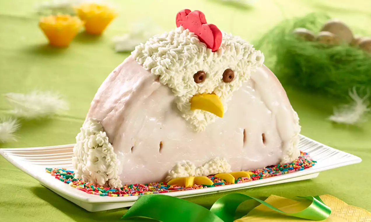 Gâteau en forme de poule blanche avec crête rouge, bec jaune, yeux en chocolat et bordé de vermicelle coloré sur un plat blanc