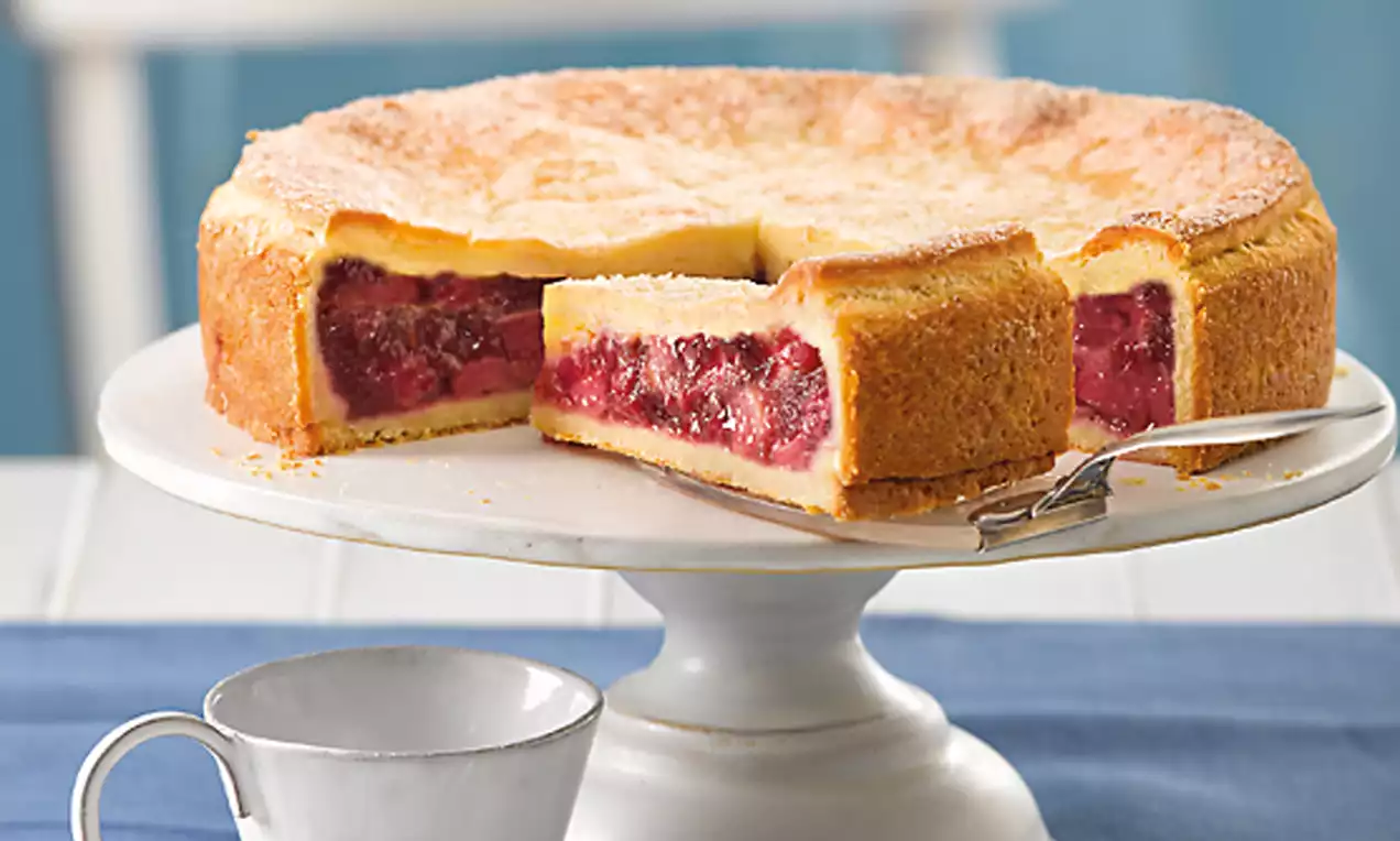 Rhabarber-Kuchen mit goldgelber, luftiger Kruste und saftig roter Füllung auf weißer Tortenplatte, daneben eine weiße Tasse auf blauem Stoff