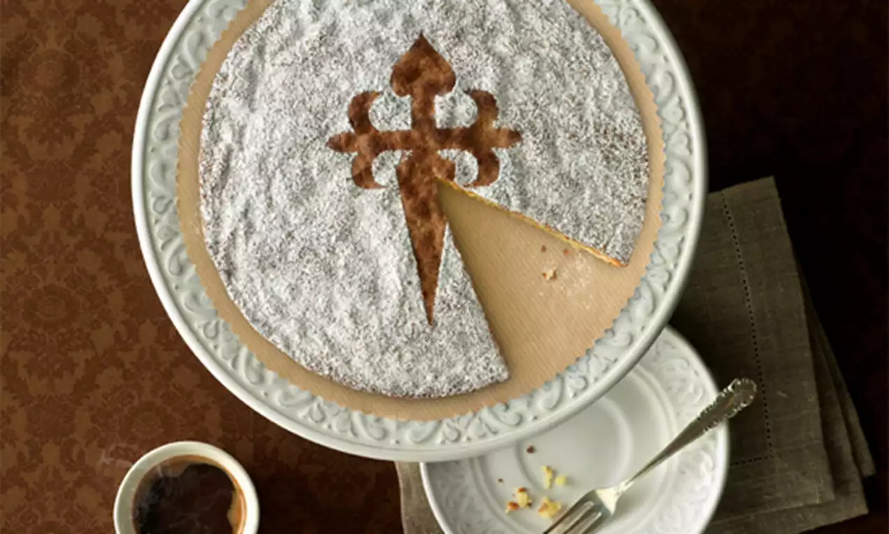 Tarta redonda de almendra cubierta de azúcar glas blanca con cruz central marrón, presentada en un plato blanco sobre mantel marrón