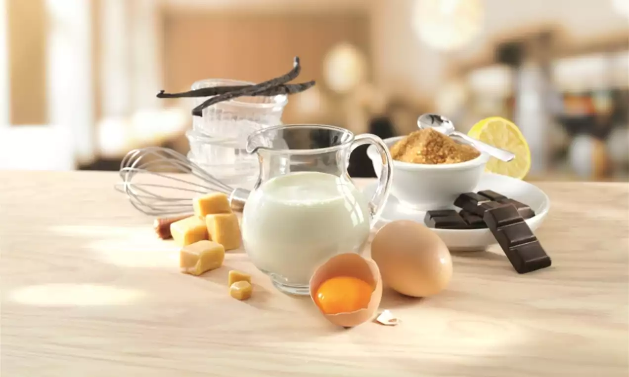 Pichet en verre rempli de lait entouré d’œufs, cubes de beurre jaune pâle, bâtons de vanille, chocolat noir et sucre brun sur une table claire