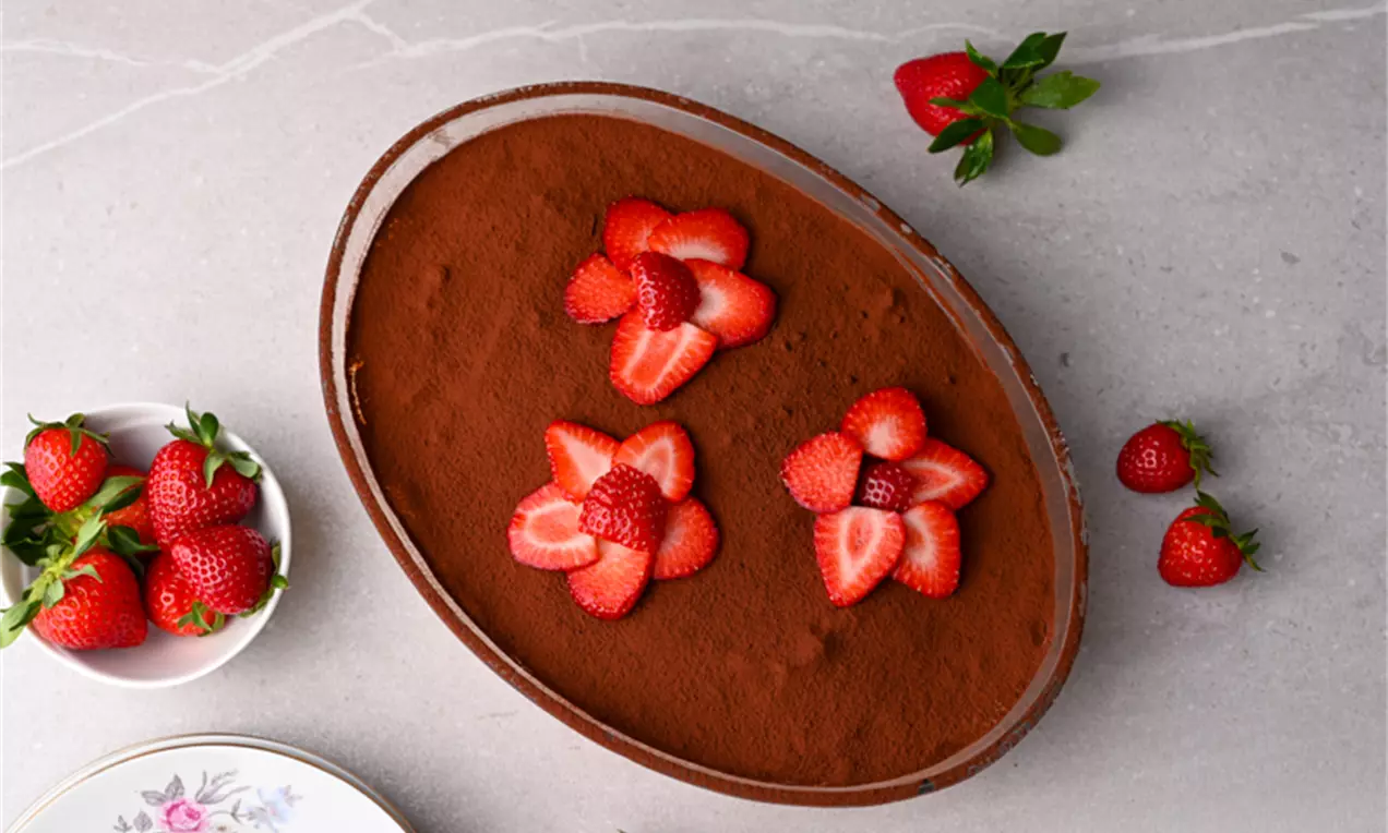 Тирамису с равномерна какаова повърхност, украсено с яркочервени резени ягоди във форма на цветя, сервирано в овална чиния