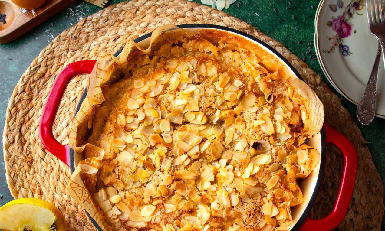 Ябълков сладкиш с топъл златист цвят, покрит с препечени филирани бадеми, в кръгла червена форма върху плетена подложка