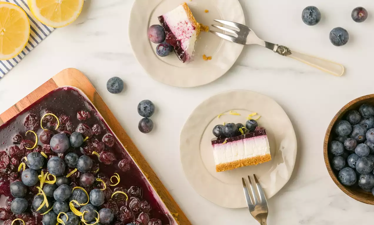 Tarta de queso con base dorada de galleta, cubierta de arándanos brillantes y ralladura de limón sobre un plato blanco con pequeños arándanos alrededor