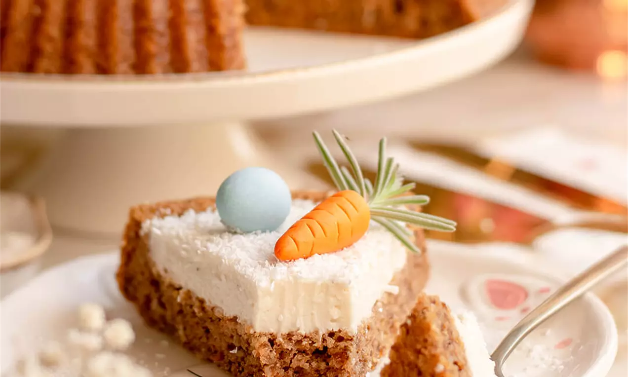 Kriška smeđe torte s bijelim kremom, ukrašena malom narančastom mrkvom, zelenim listićima i pastelno plavim jajetom na tanjuru s motivom zeca