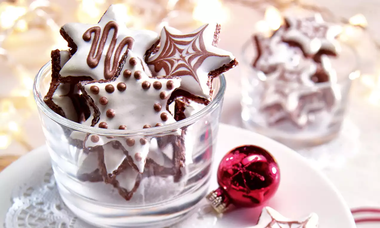 Zvjezdasti keksi smeđe boje s bijelom glazurom i čokoladnim ukrasima u staklenoj zdjelici, uz crvenu ukrasnu kuglicu na bijelom tanjuru