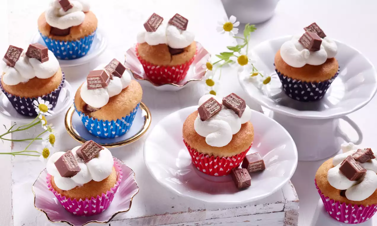 Goldene Mini-Muffins in bunten Papierförmchen, mit fluffiger Sahnehaube und Schokoladenwürfeln verziert, auf weißen Tellern mit Blumen dekoriert