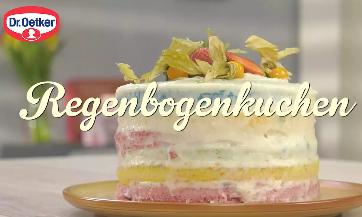 Vielschichtiger Regenbogenkuchen mit farbigen Teigschichten, heller Creme und dekorativen Früchten auf einem braunen Teller drapiert