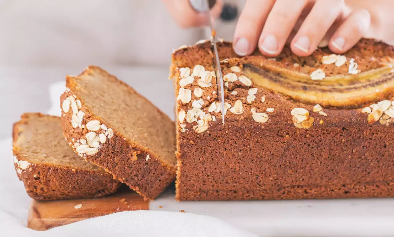 Plum cake rectangular dorado con interior esponjoso y corteza decorada con rodajas de plátano y copos de avena sobre una tabla blanca