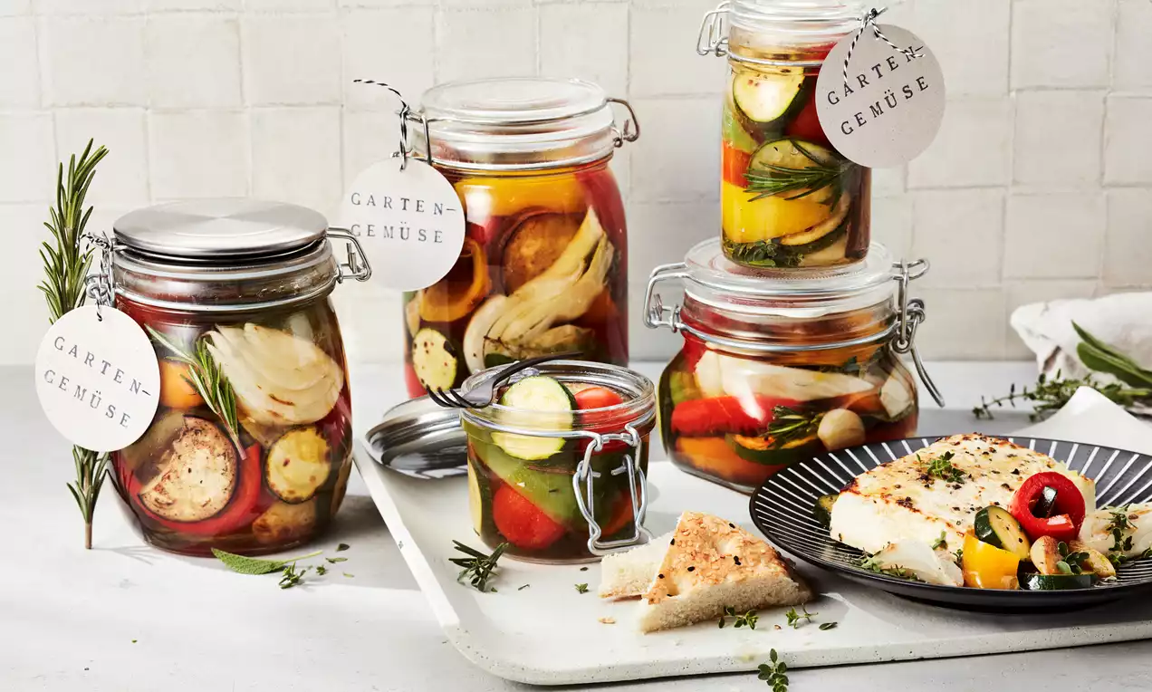 Eingelegtes Gartengemüse in bunten Schichten mit Zucchini, Paprika und Kräutern in Gläsern mit Metallbügelverschluss auf weißem Tablett