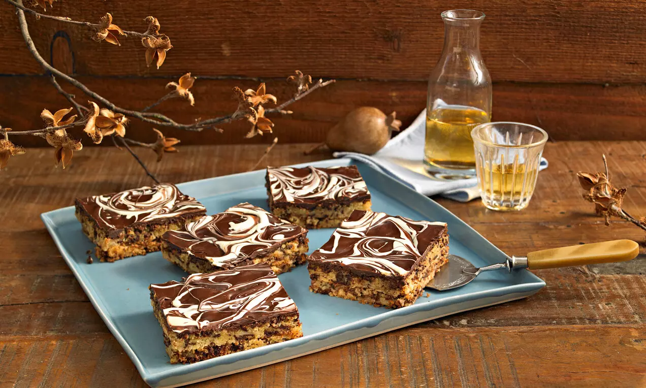 Нарязани квадратни парчета мраморен сладкиш с тъмнокафява глазура, върху светлосиня чиния, декорирана със свежи клонки и поставена на дървен фон