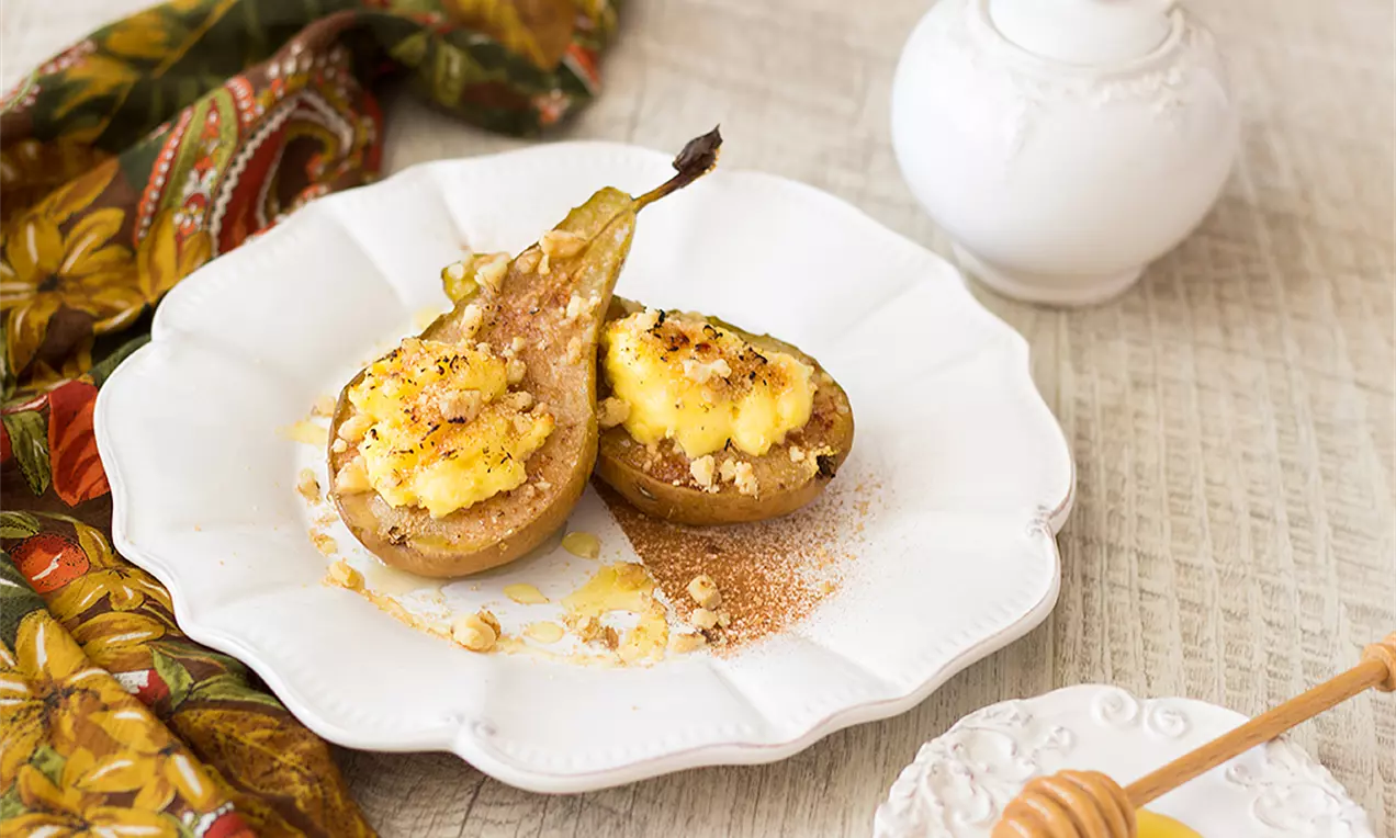 Златисто-кафяви половинки печени круши с обгарен крем Брюле, поръсени с трохи и канела, подредени върху бяла чиния с флорални детайли