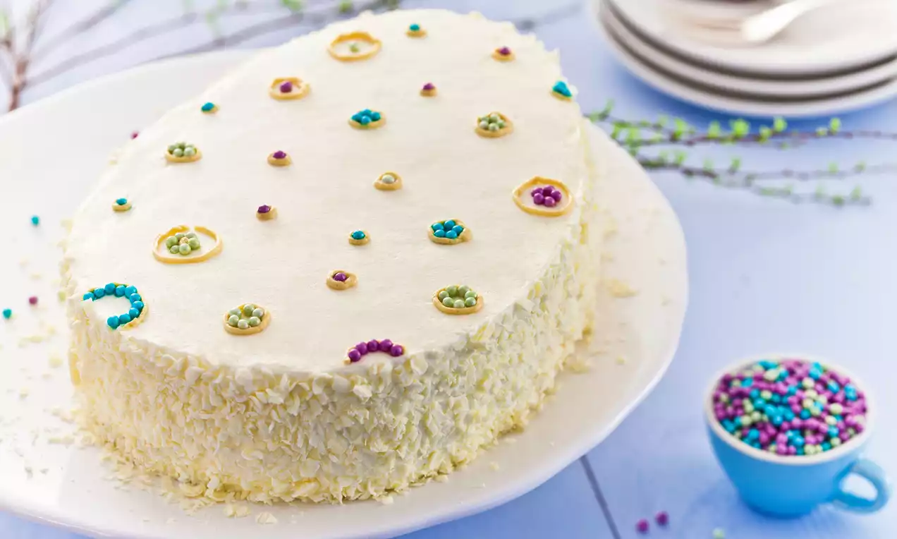 Ovale Sahnetorte mit weißer Creme, bestreut mit bunten Perlen und gelben Zuckerringen, auf einer weißen Platte mit Streudekor
