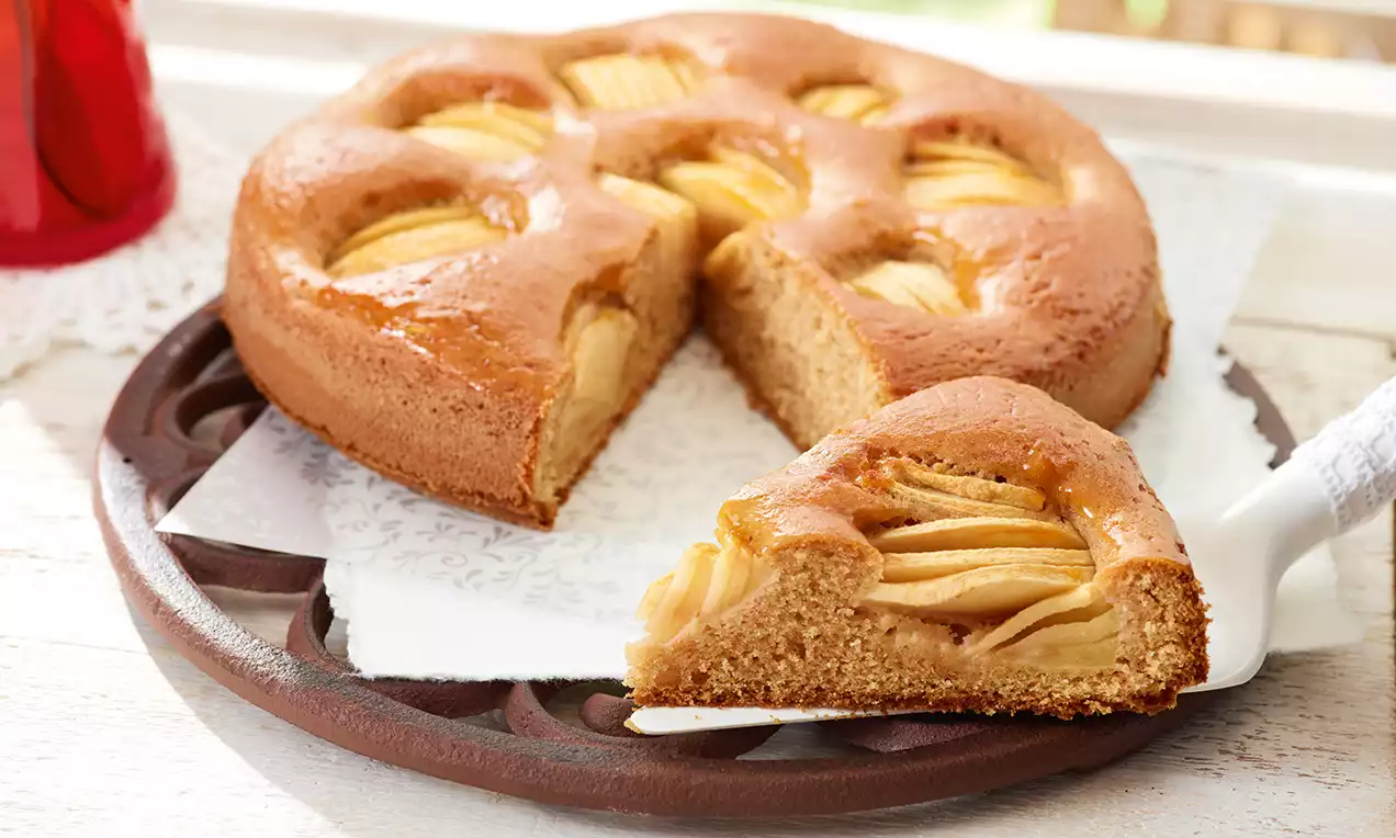 Rund gebackener Vollkornkuchen mit goldenen Apfelscheiben in weicher Teigkruste auf brauner Platte mit ausgeschnittenem Stück auf weißer Serviette