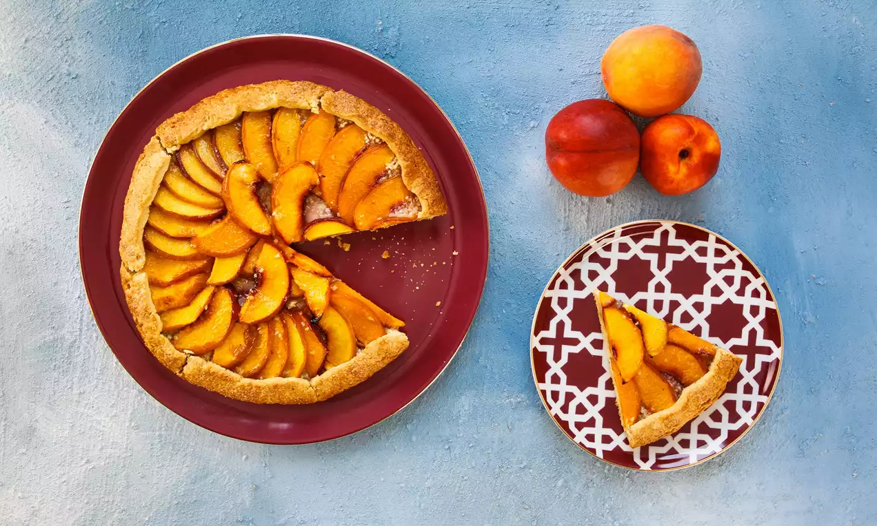 Altın rengi ince bir hamurun üzerinde dilimlenmiş şeftalilerle bezelmiş galette, kırmızı düz bir tabakta ve bir dilimi desenli bir tabakta