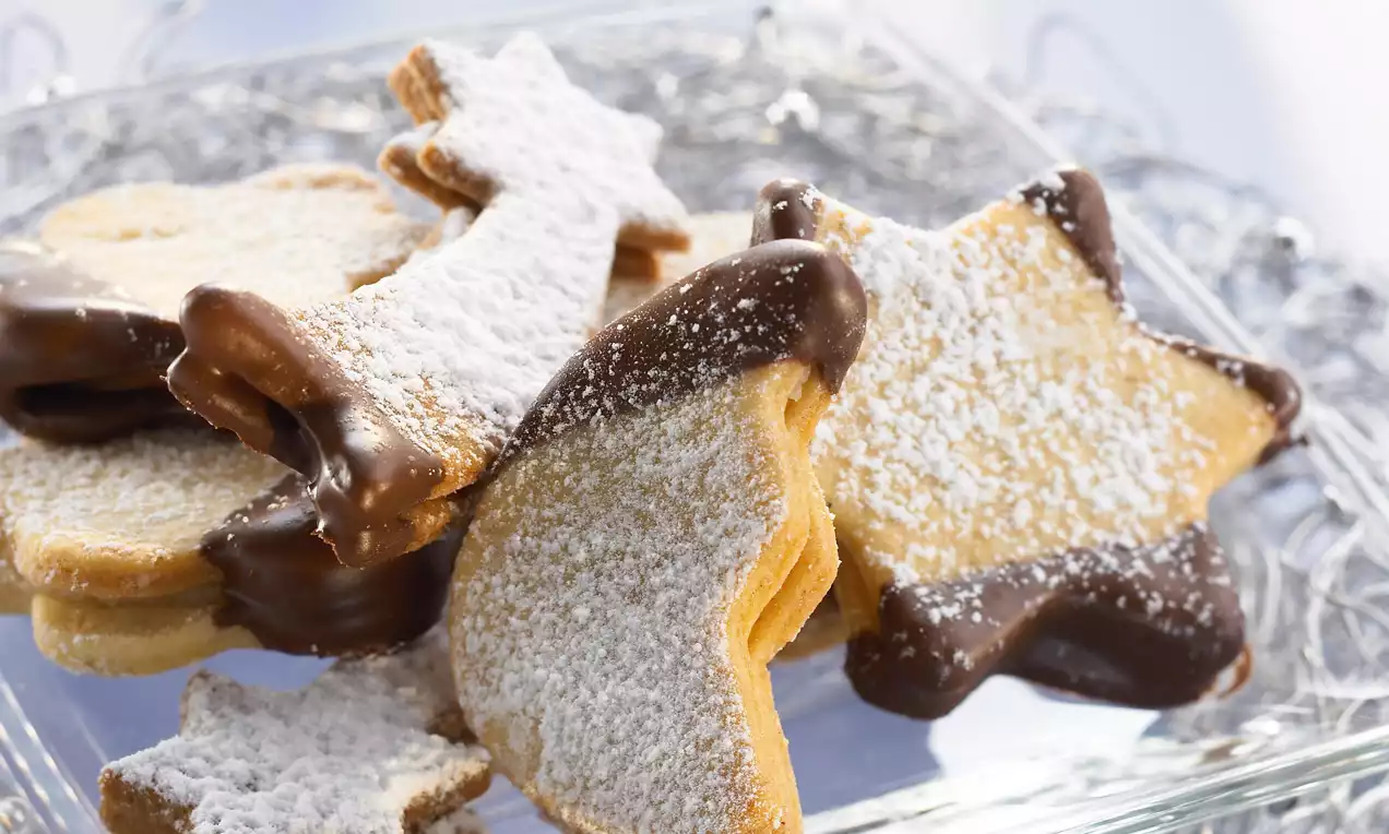 Goldgelbe Stern- und Mondförmige Butterguetzli mit glänzendem Schokoladenrand und feinem Puderzucker auf einer Glasplatte angerichtet