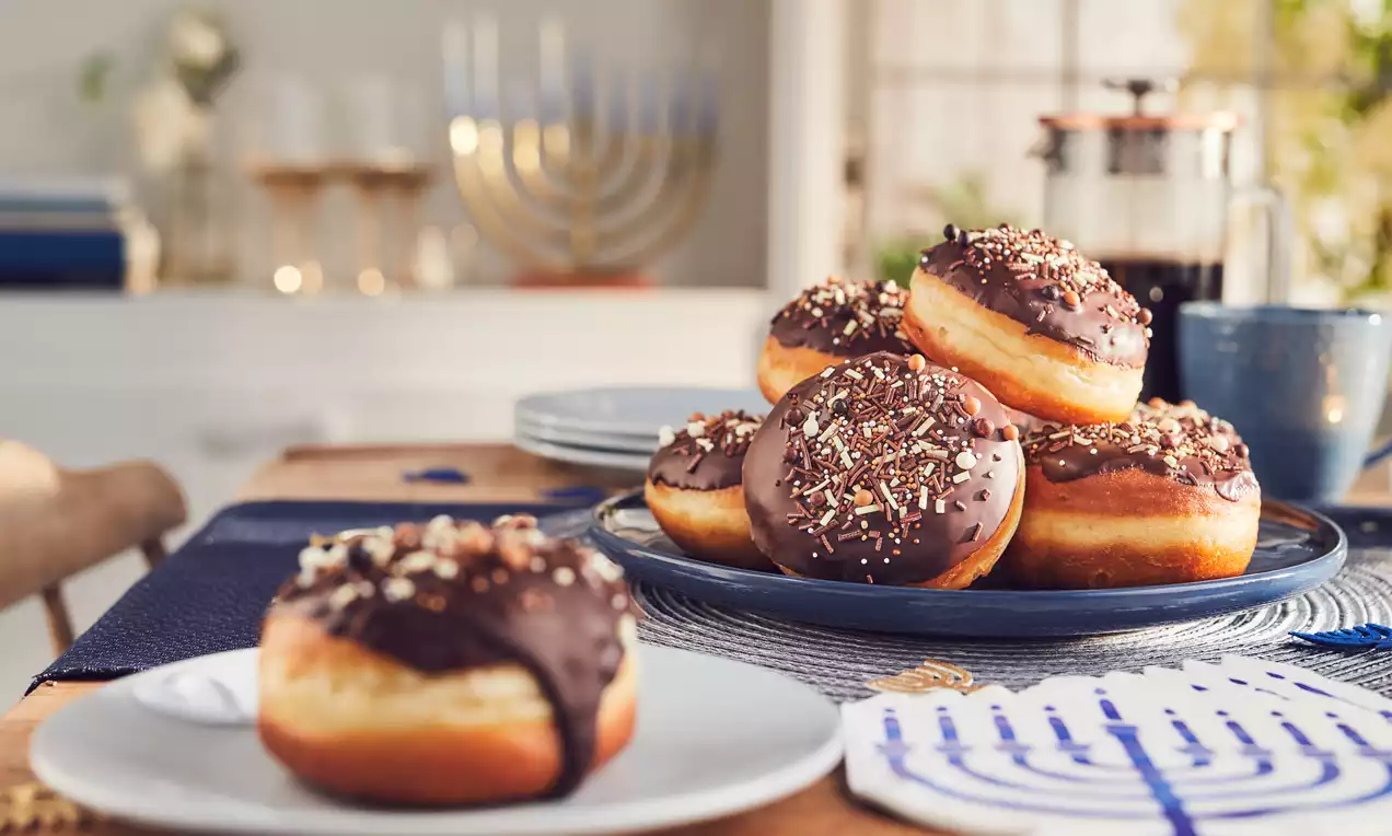Golden, round sufganiyot topped with glossy dark chocolate and colourful sprinkles, arranged on plates with festive table decor