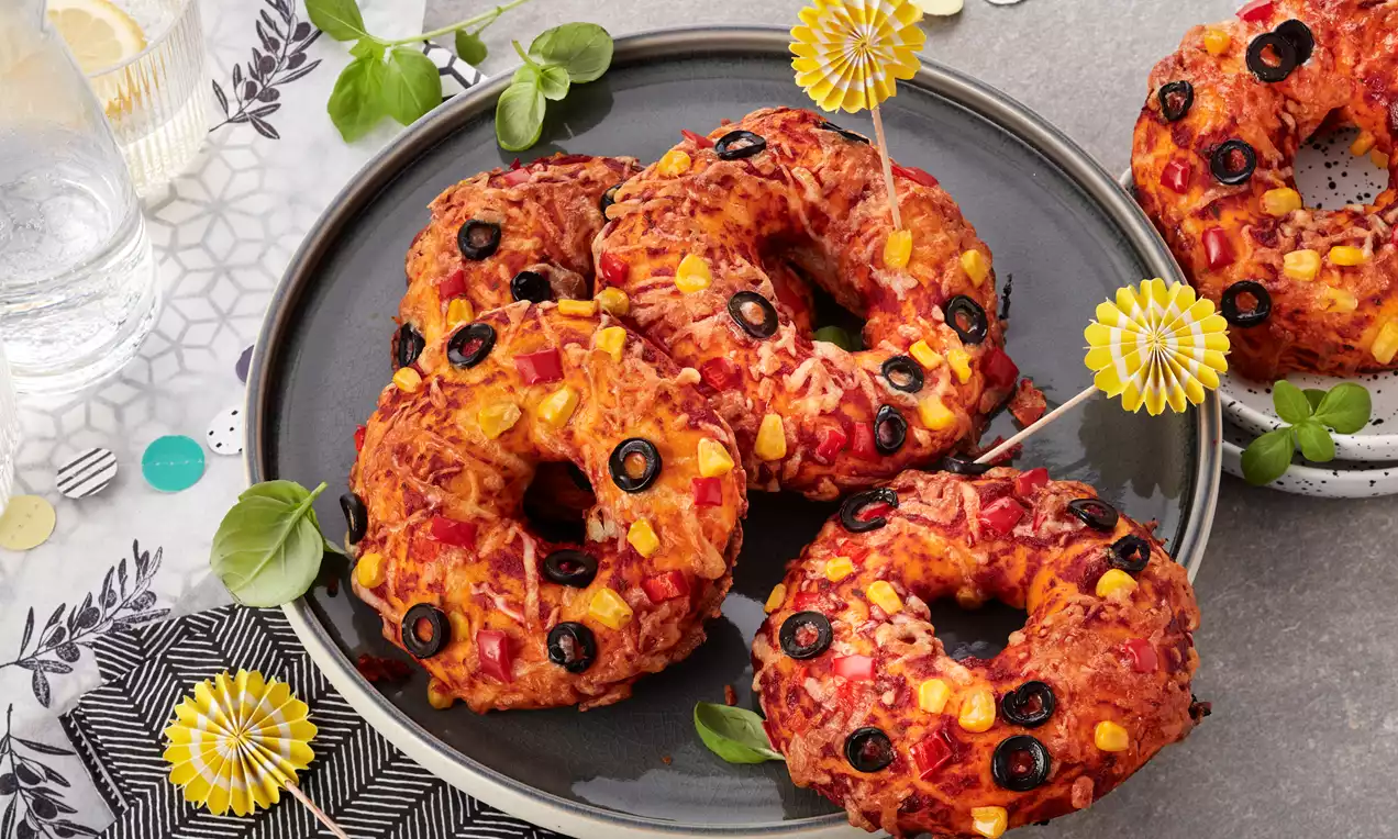 Goldbraune Pizza-Donuts mit geschmolzenem Käse, gelbem Mais und schwarzen Olivenscheiben dekorativ auf einem Teller mit Basilikum-Blättern