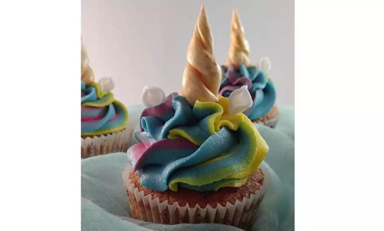 Cupcake surmonté d’un tourbillon de crème arc-en-ciel pastel et d’une corne dorée torsadée avec de petites décorations blanches