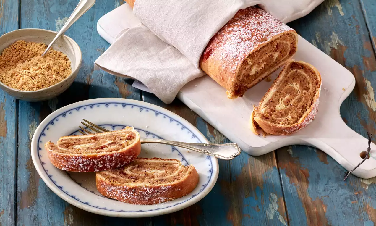 Goldbrauner Nussstrudel mit feiner Füllung, zart mit Puderzucker bestäubt, auf Holzbrett und Teller mit Besteck und Schälchen dekoriert