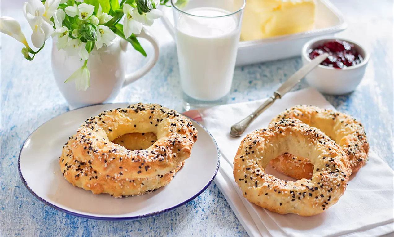 Златисти гевречета с поръска от черен и бял сусам, кръгли и пухкави, подредени върху бяла чиния до чаша мляко и свежа декорация с цветя