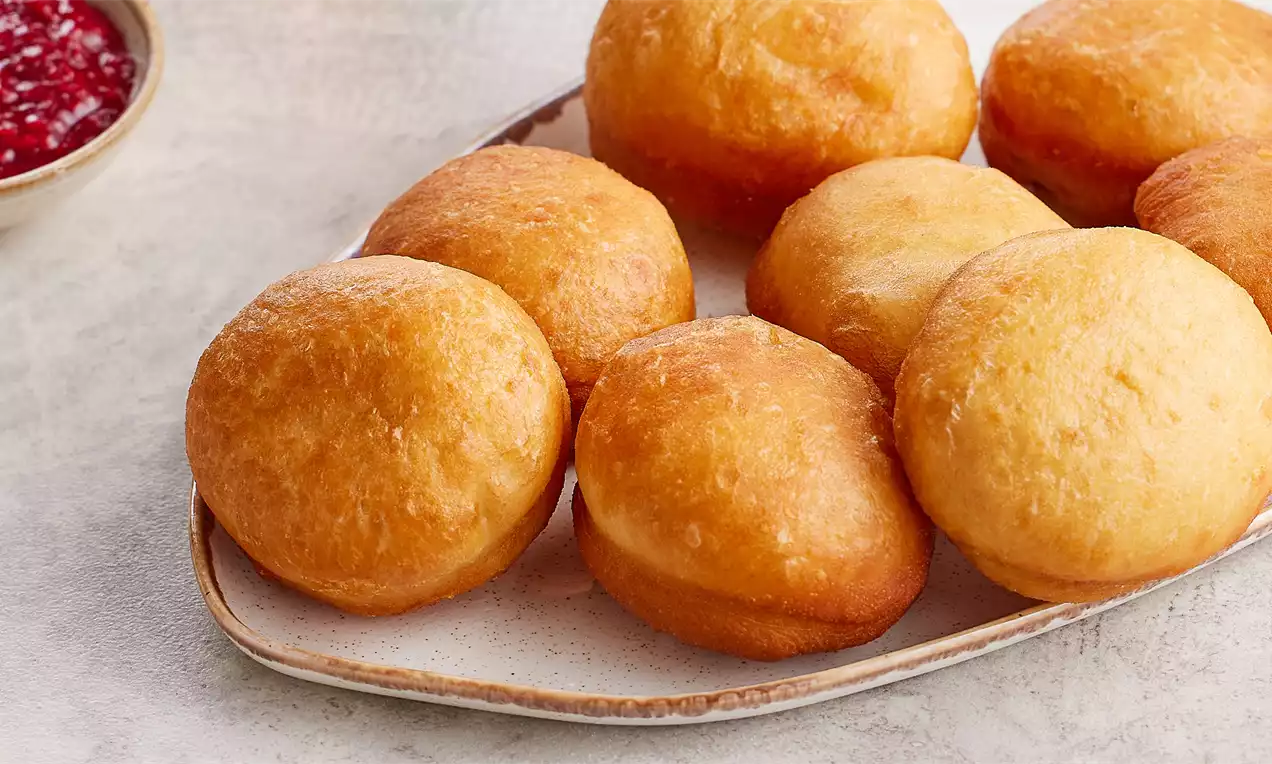 Altın rengi ve yuvarlak kızarmış hamur işleri, krem rengi bir tabakta düzenli şekilde yerleştirilmiş, yanında sos kâsesi bulunuyor