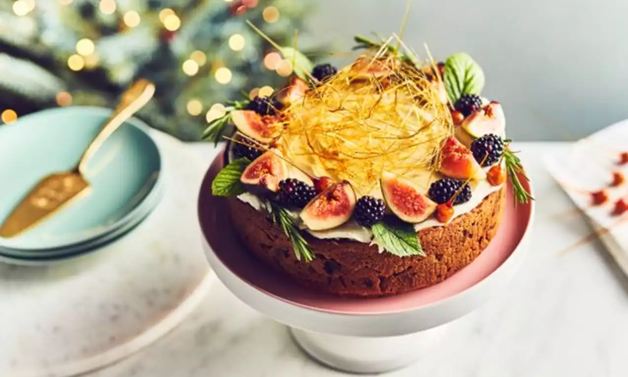Golden fig cake with a textured surface, topped with fresh figs, blackberries, mint leaves, spun sugar and white cream on a pink pedestal