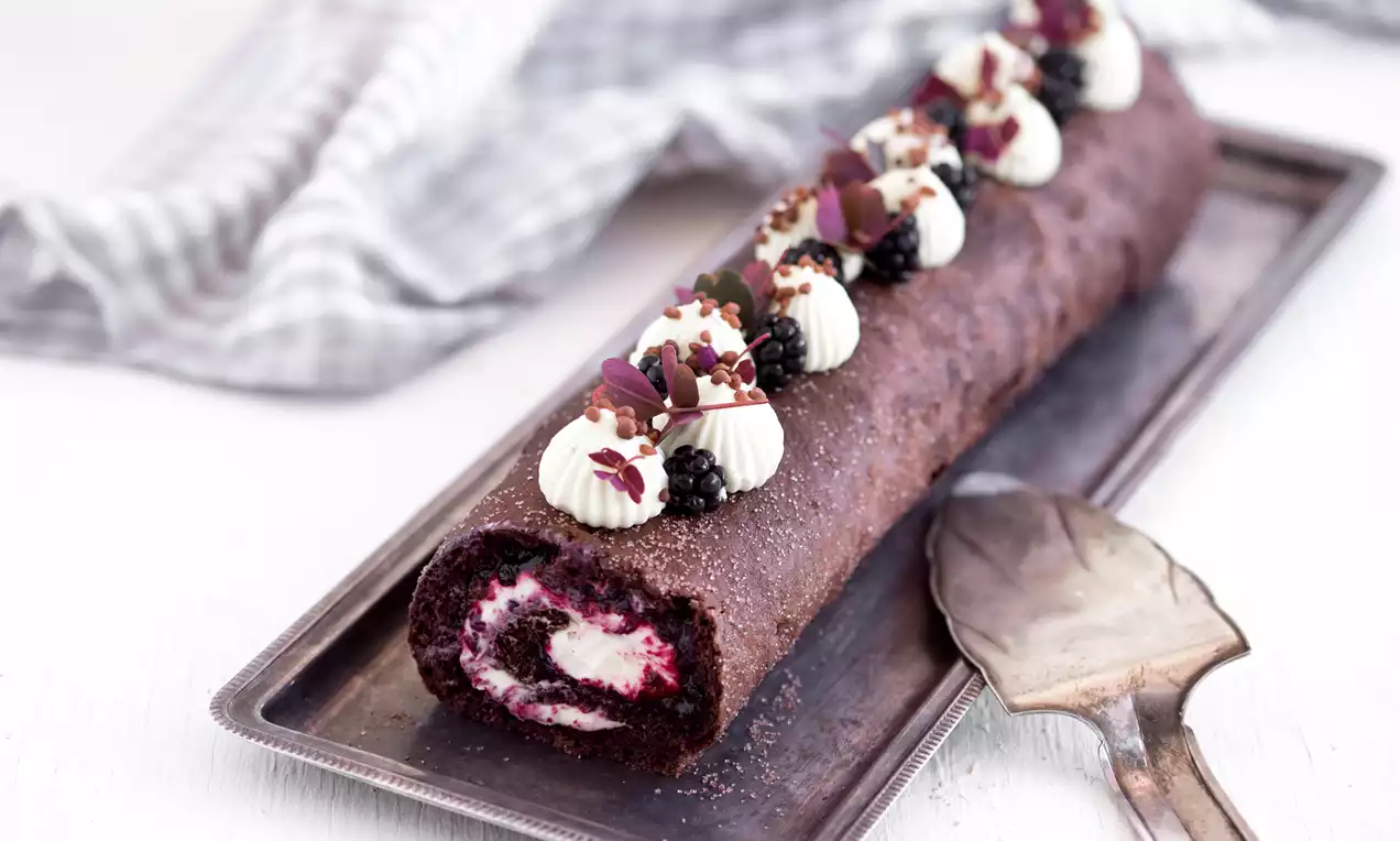 Aflang chokoladebrun roulade med hvid fyld og brombær, pyntet med fløde, blade og crumble på et sølvfad ved siden af en serverske
