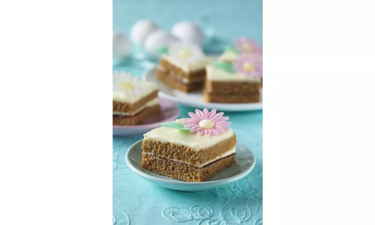 Perníkové řezy s bílou vrstvou krému, zdobené barevnými jedlými květinami, na malých talířcích na modrém ubrusu s jemným vzorem