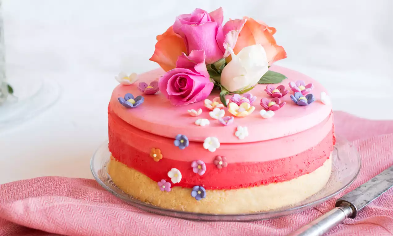 Runde Torte mit rosa Farbverlauf auf Keksboden, dekoriert mit bunten Zuckerblüten und echten Rosen auf Glasplatte mit rosa Stoff drapiert