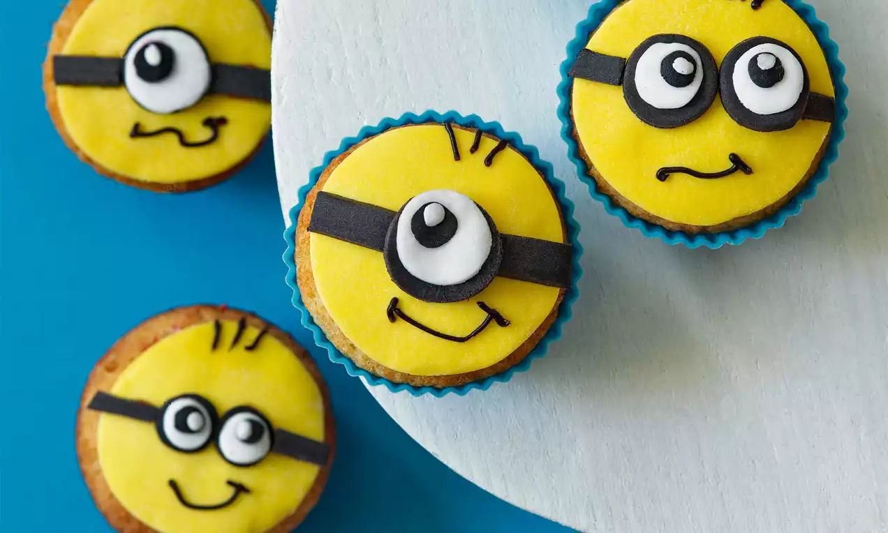 Banánové muffiny s kulatou žlutou polevou zdobené černými pásky, bílými očky a kreslenými detaily, uspořádané na modrém podkladu