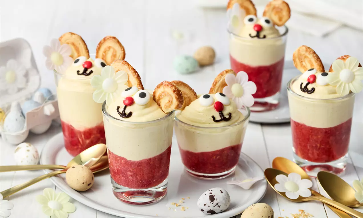 Osterdessert im Glas mit gelber Creme, roten Früchten, Ohren aus Blätterteig, Zuckeraugen und Blumen-Deko auf weißem Teller mit Eiern